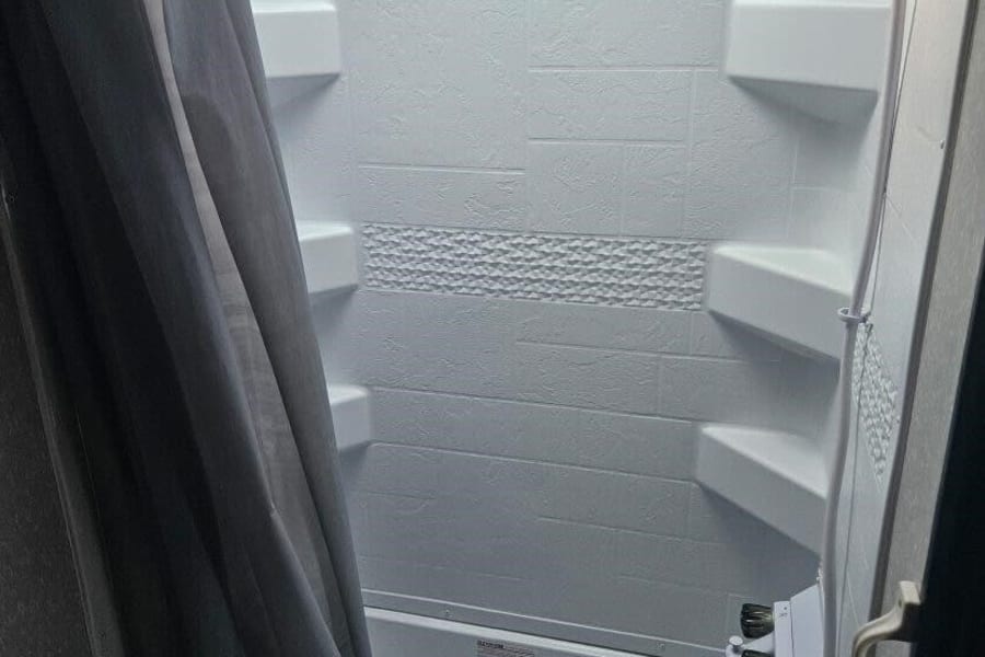 Compact RV rental bathroom featuring a white shower/tub, textured walls with shelves, and a gray shower curtain.