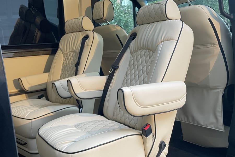 Luxurious RV rental interior: cream-colored captain's chairs with diamond stitching, black piping, and an overhead screen displaying news.