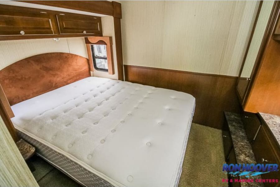 RV rental bedroom featuring a queen bed, wooden headboard, overhead cabinets, and a side window for a comfortable stay.