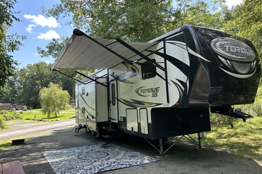 A spacious Torque SS fifth-wheel rv rental is parked at a tranquil, shaded campsite. Awning extended over a comfy rug.