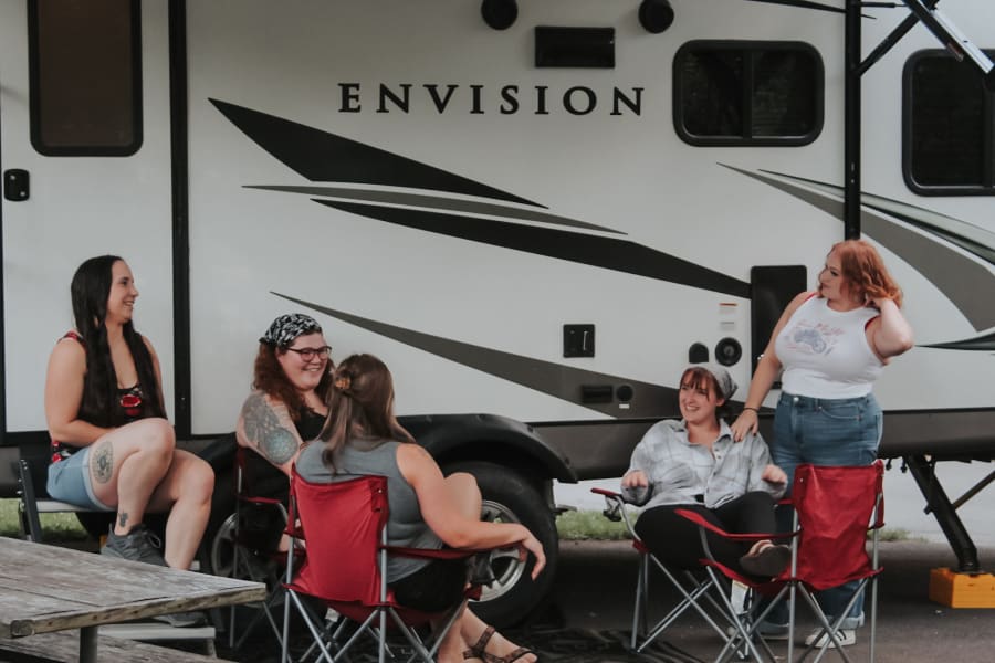 Group of women relaxing, chatting, and laughing by their white Envision RV rental at a campsite.