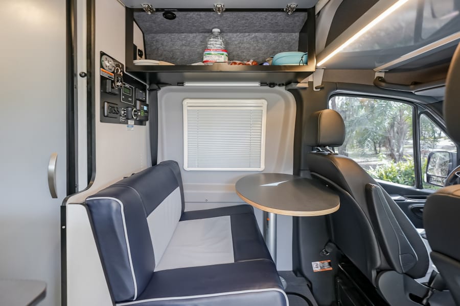RV rental interior shows blue/white dinette, adjustable table, control panel, and overhead storage, designed for efficient travel.