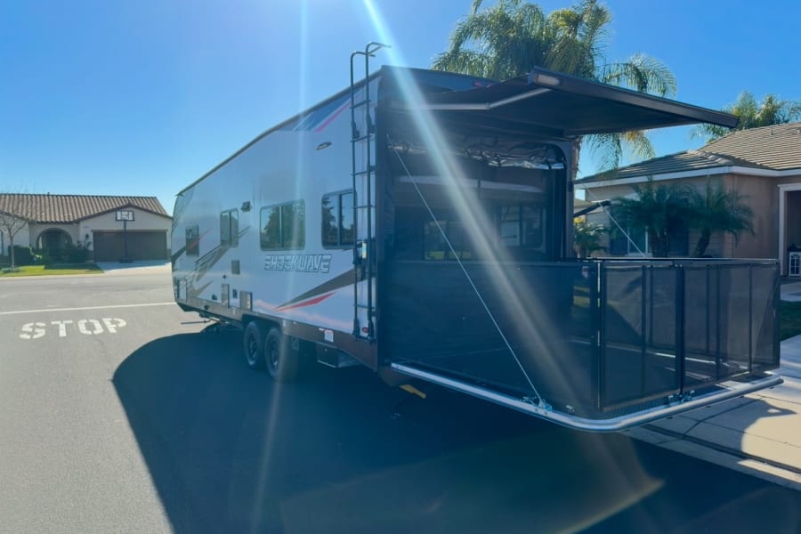 Toy hauler RV rental: Rear ramp lowered, creating a patio with safety rails and awning, parked on a sunny street.