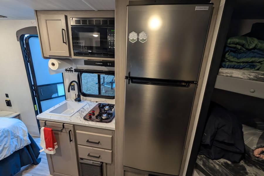 RV rental interior with a compact kitchen (fridge, stove, microwave), a main bed, and bunk beds for travelers.
