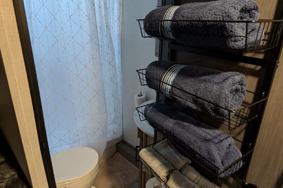 Compact RV rental bathroom with a white toilet, shower curtain, and wall-mounted rack holding neatly rolled towels and toilet paper.