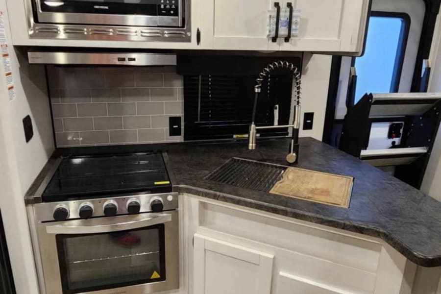 This RV rental features a well-equipped, compact kitchen with stainless steel microwave, oven, range, and sink, set against white cabinetry and dark counters.
