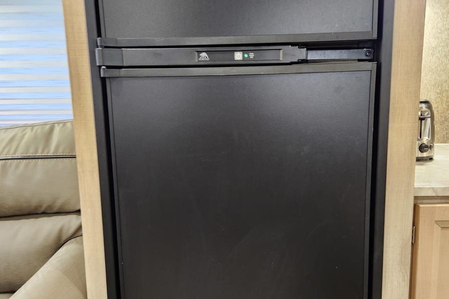 Modern black RV refrigerator/freezer in an RV rental kitchen, integrated into light wood cabinetry. Shows control panel with green light.