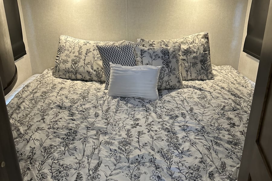 This comfortable RV rental bedroom features a large bed with black and white floral bedding, decorative pillows, and reading lights.