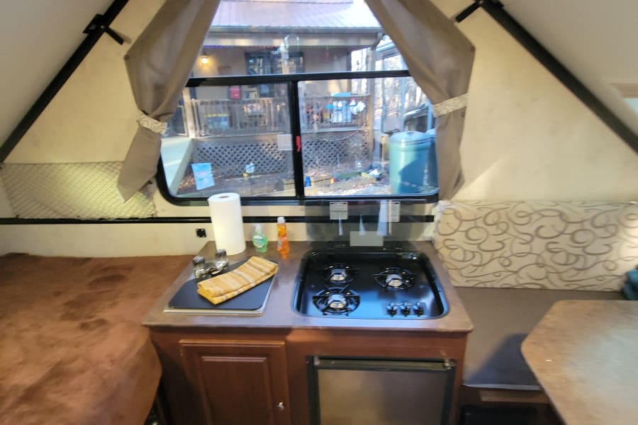 RV rental interior showing a compact kitchen with stove/sink, bed, dining area, and exterior view through the window.