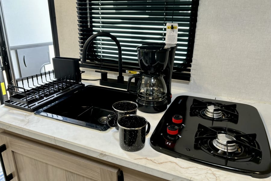 Well-equipped RV rental kitchen featuring a black sink, two-burner stove, coffee maker, and camp mugs on a light counter.