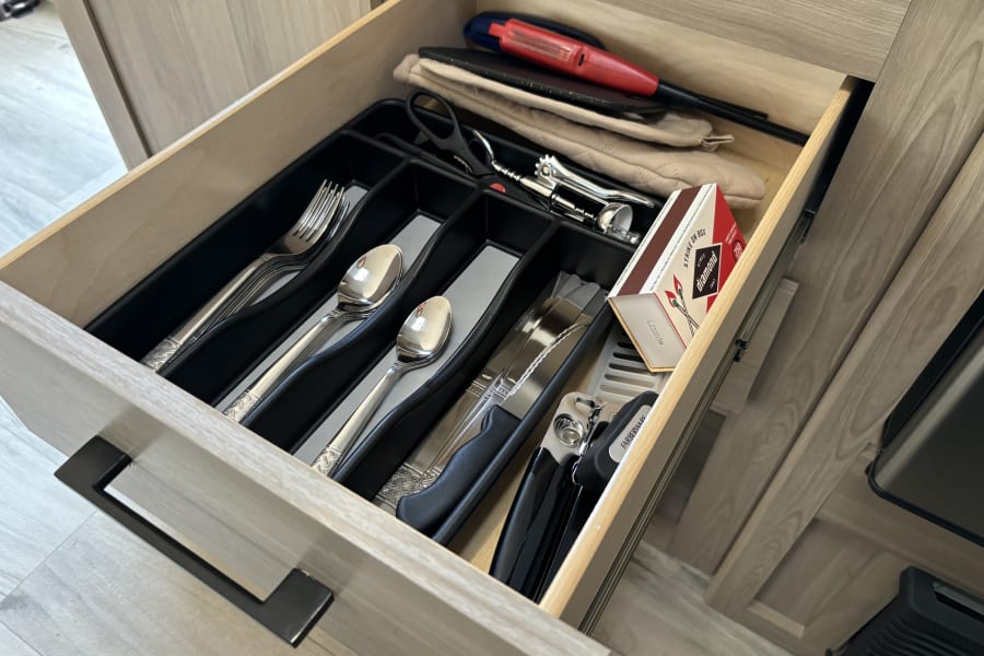 Open RV rental drawer revealing organized cutlery, oven mitts, matches, and kitchen gadgets for everyday use.