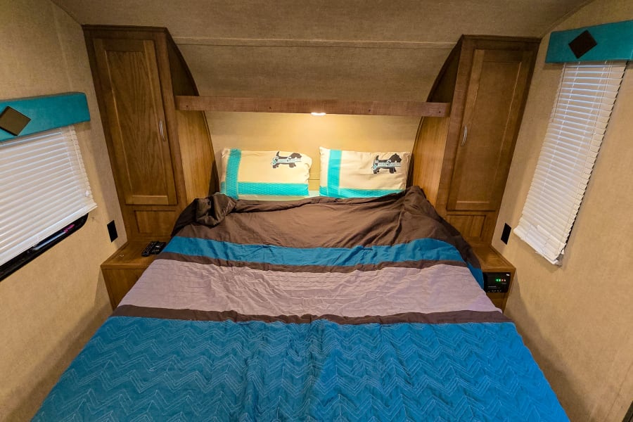 This RV rental interior shows a cozy bedroom with a large bed, brown and teal bedding, dog pillows, and wooden cabinets.
