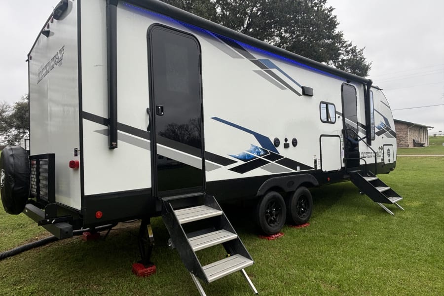 Modern travel trailer RV rental, featuring blue awning lights and sturdy steps, parked on green grass.