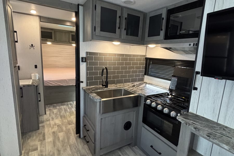 Modern RV rental interior: kitchen with grey subway tile, stainless sink, stove, microwave, and glimpse of bedroom.