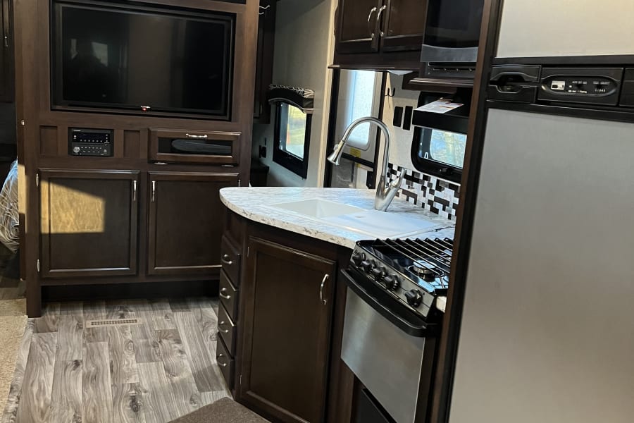 Modern rv rental interior: compact kitchen with dark wood cabinets, sink, stove, fridge, and a living area with TV.
