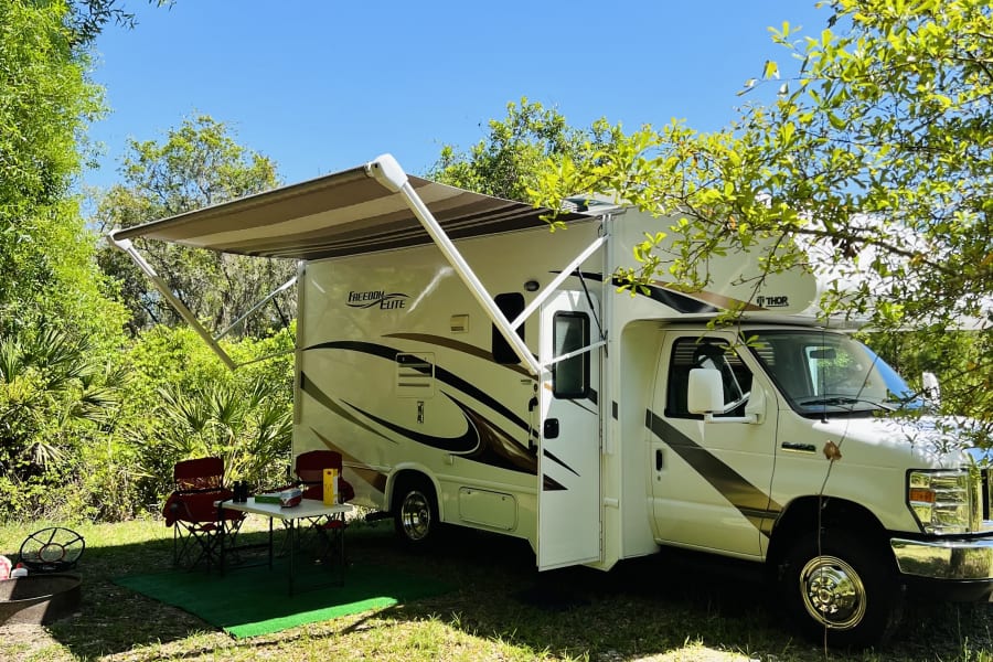 White Class C RV rental with awning extended at a sunny green campsite. Chairs, table, and fire pit are ready amidst lush trees.