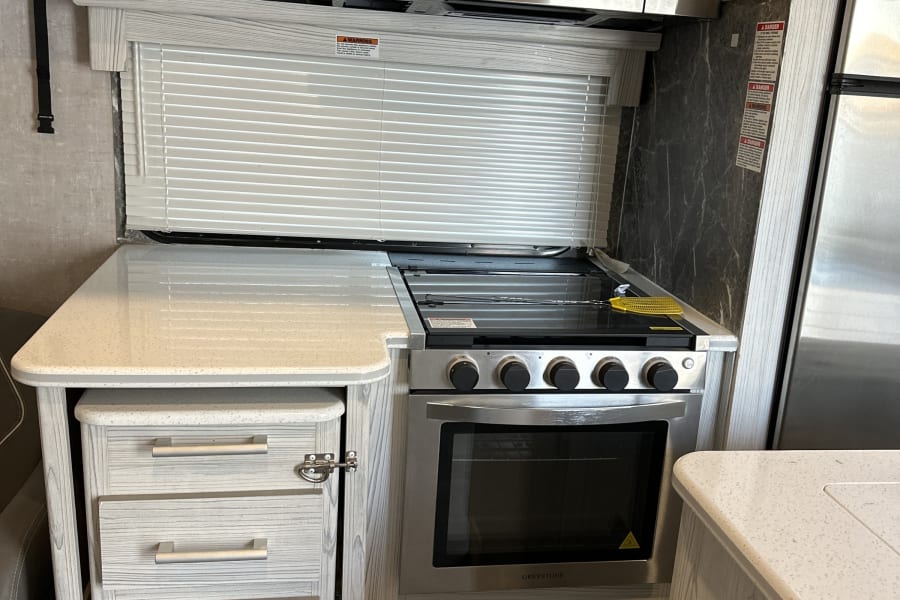 RV rental kitchen featuring a GE microwave, Greystone stove, light wood cabinets, speckled counters, and window blinds.
