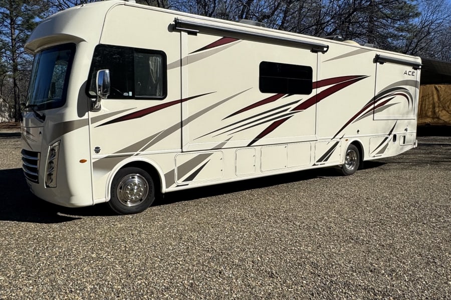 Cream-colored RV rental with maroon and gray stripes parked on a sunny gravel lot, surrounded by trees.
