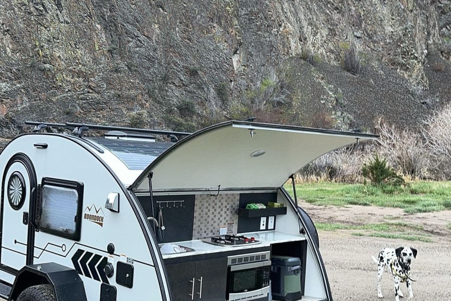 Teardrop RV rental with an open outdoor kitchen parked by rocky mountains. A Dalmatian dog walks on grass. Ideal for camping.