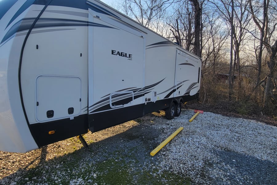 Experience outdoor living! This spacious white fifth-wheel RV rental is parked on a gravel lot amidst bare trees.