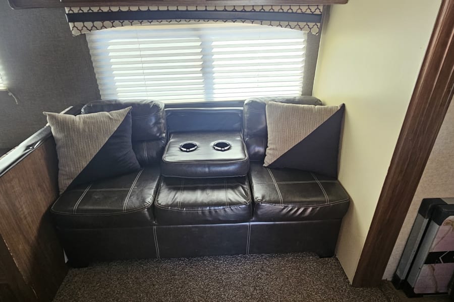 Relax in this RV rental's cozy interior, featuring a dark leather sofa with cup holders, pillows, and window blinds.