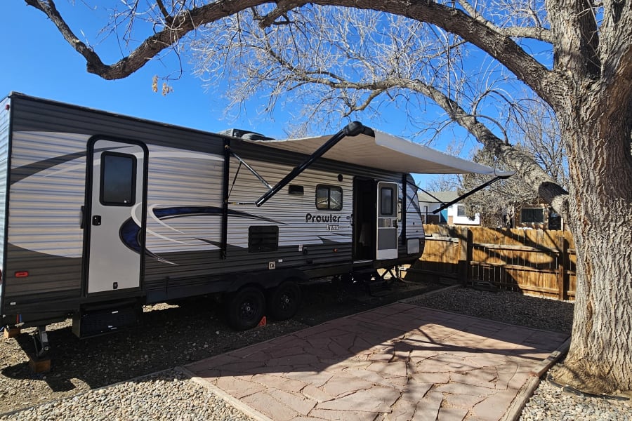 This Prowler Lynx travel trailer RV rental is parked on a gravel lot with its awning extended beside a patio.