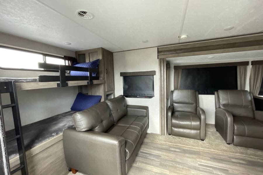 This RV rental's interior shows a living space with a gray sofa, two recliners, and bunk beds with blue pillows.
