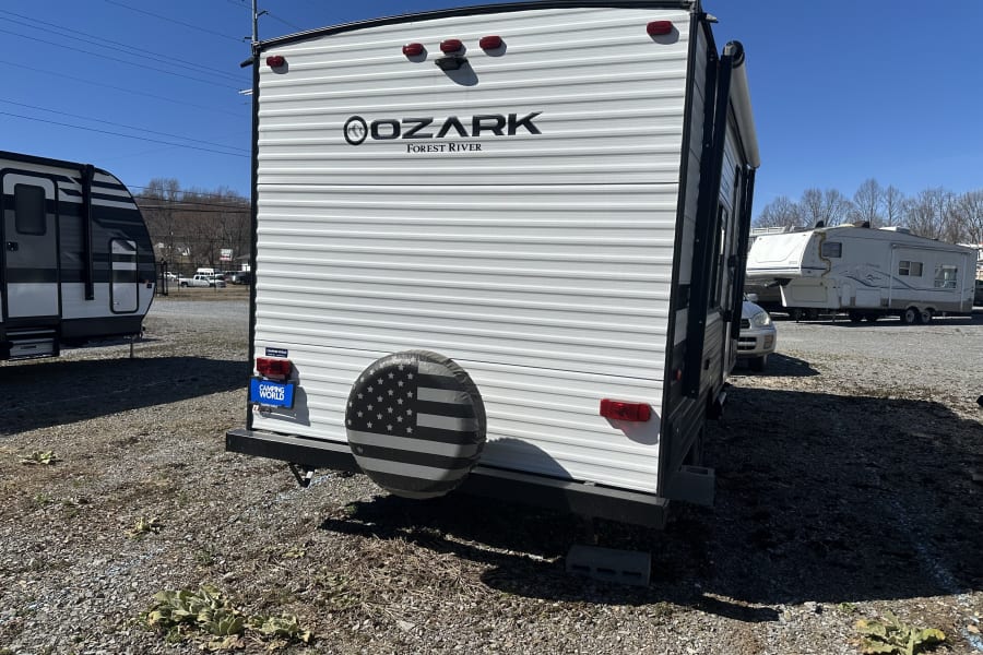 Rear view of an Ozark Forest River travel trailer (RV rental) on a gravel lot. Features a grayscale American flag tire cover.