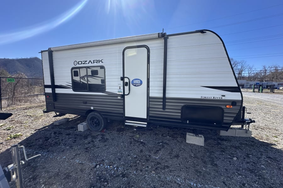 A white and gray pre-owned Forest River Ozark travel trailer for an RV rental, parked on gravel under a sunny sky.