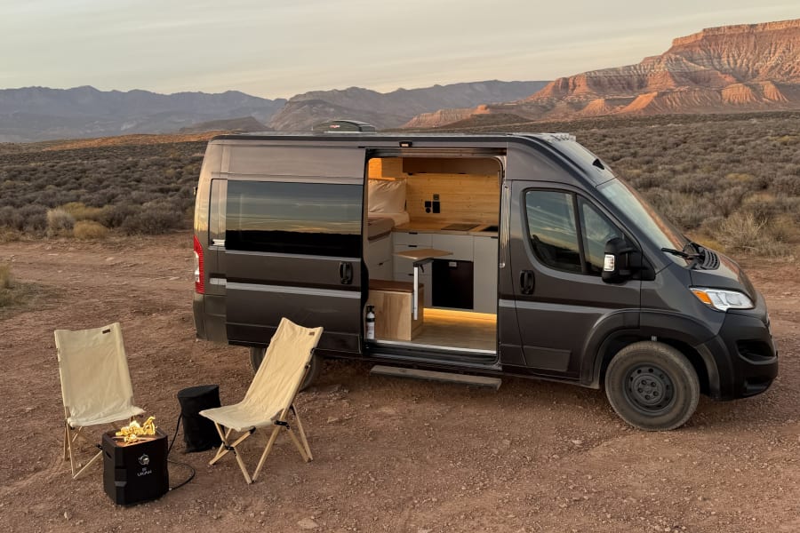 An RV rental camper van, side door open to its lit interior, with chairs and a fire pit, parked in a desert landscape.