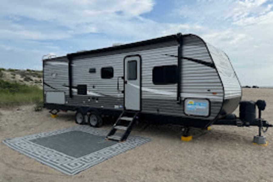 Your next rv rental adventure awaits! A modern silver & grey travel trailer is parked on a sandy beach with an outdoor rug & steps.