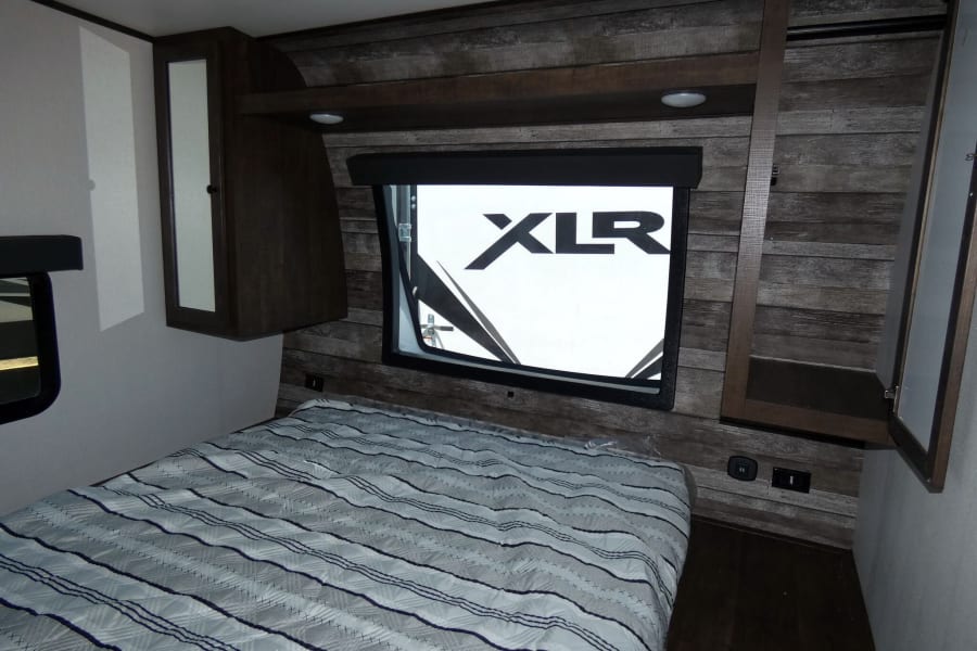RV rental interior: a compact bedroom with a cozy striped bed, window displaying 'XLR' logo, and built-in storage.