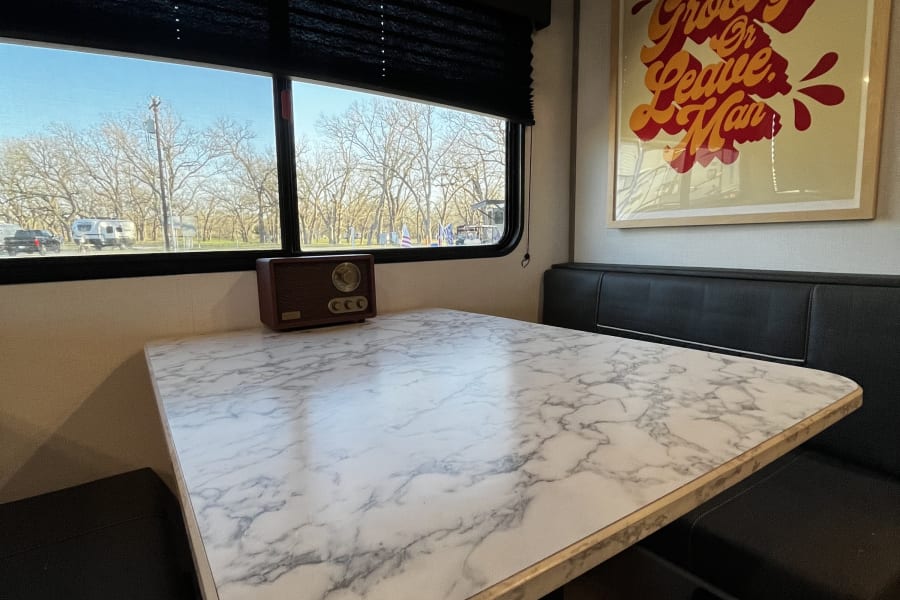 Enjoy your RV rental! A cozy dinette with a marble table, comfy seating, and a window view of a sunny campground.