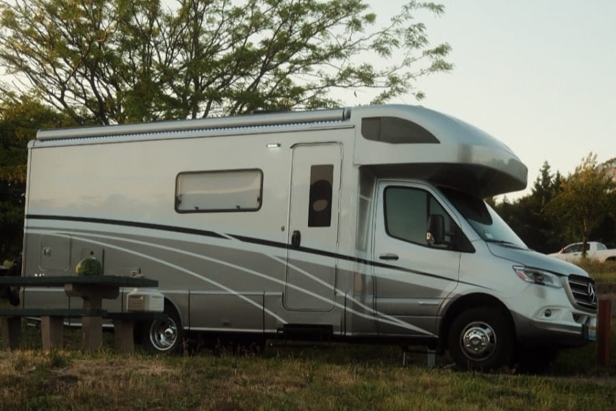 Modern silver RV rental parked at a campsite. A person sits at a picnic table with a watermelon nearby in golden hour light.
