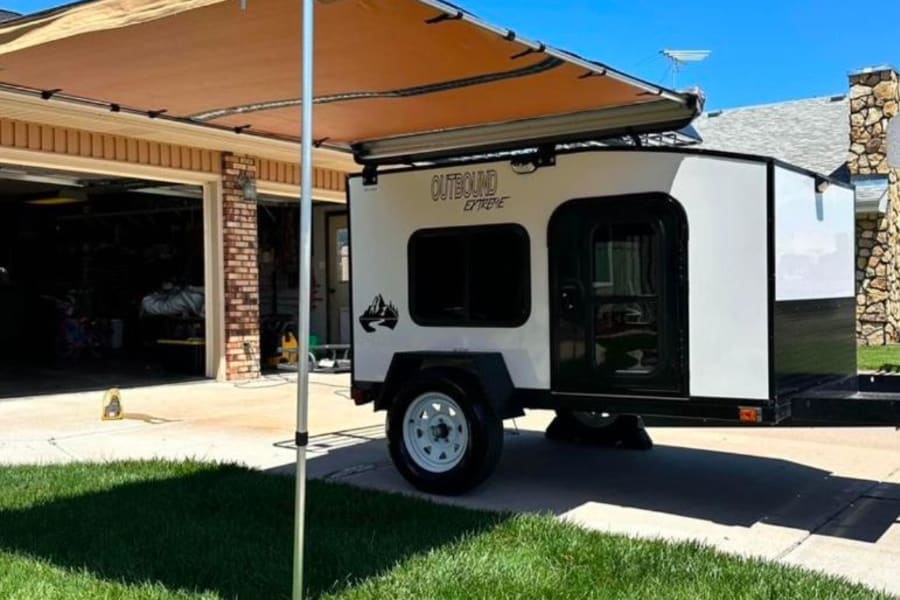 This small white and black 'Outbound Extreme' teardrop camper with an extended awning is perfect for an rv rental adventure.