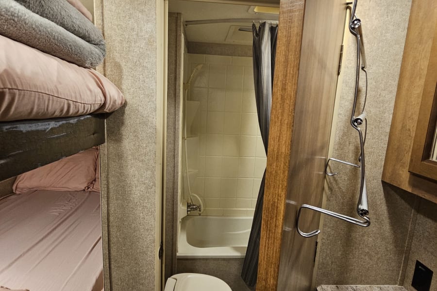 An RV rental interior showing a compact bathroom with a toilet and shower, bunk beds, and a sink area.