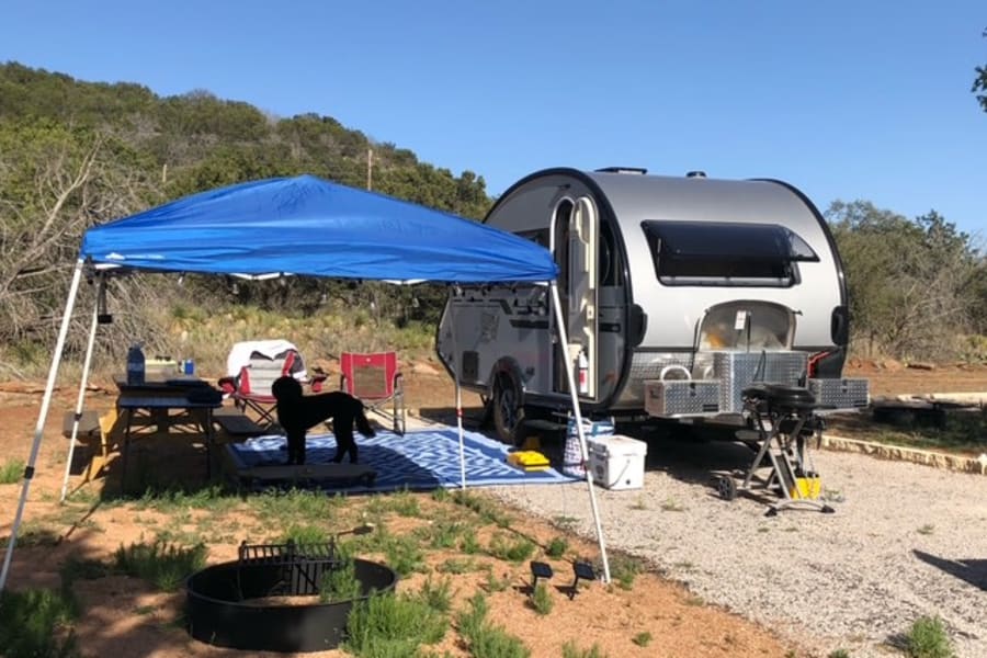 RV rental campsite setup with a teardrop trailer, blue canopy, dog, chairs, and grill for outdoor living.