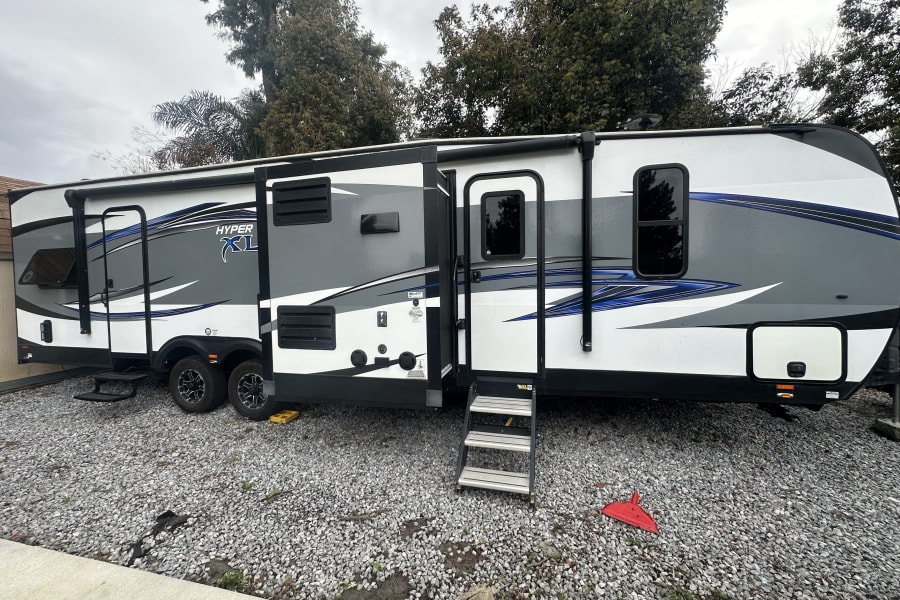 Spacious RV rental, a white, gray, and blue travel trailer, is set up on a gravel lot under cloudy skies.