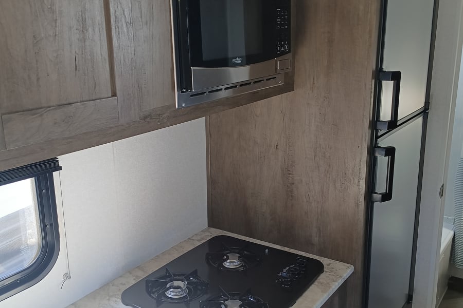 RV rental kitchen featuring a stainless steel microwave, large refrigerator, and a three-burner gas stovetop on a light counter.
