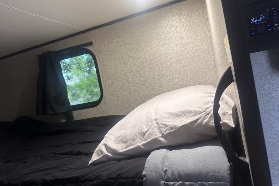 Relax in your RV rental's cozy bunk bed, with a window offering nature views and an integrated entertainment system.
