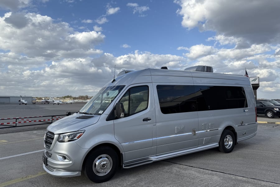 Silver Airstream RV rental parked at an airport tarmac with private jets and cloudy skies.