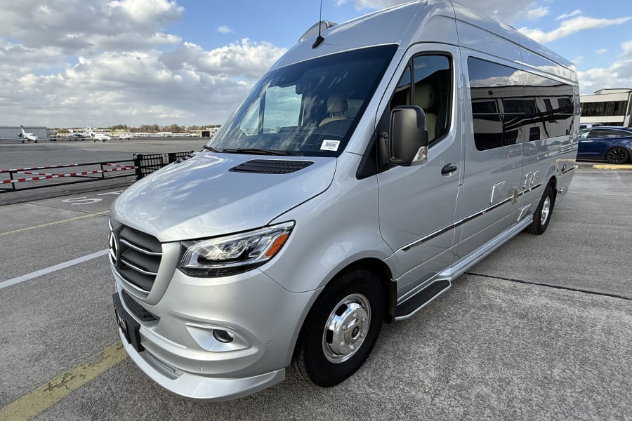 Silver Airstream RV rental, a sleek camper van, parked on airport tarmac with jets visible under a cloudy sky.
