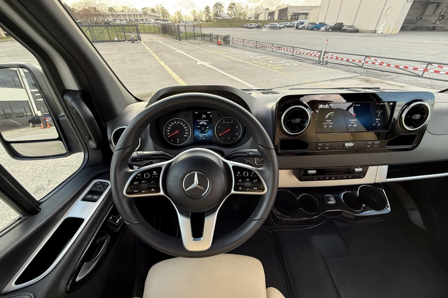 Mercedes-Benz Sprinter rv rental cockpit: dashboard, steering wheel, & driver's view of an airport with hangars and aircraft.