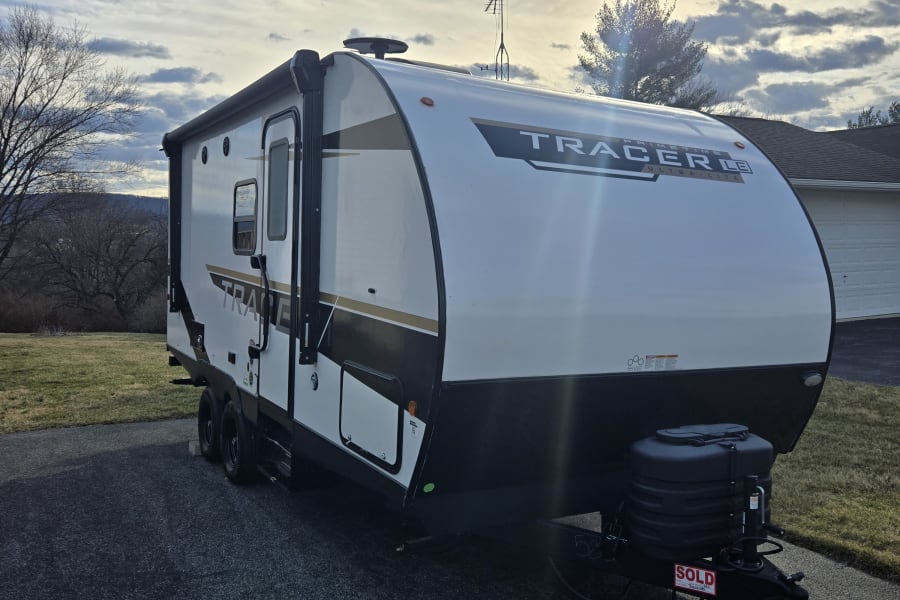 A new white Tracer travel trailer is parked on a driveway on a sunny day. Looks like a great candidate for an RV rental.