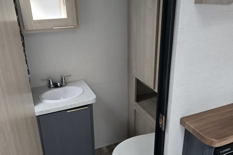 Modern compact RV rental bathroom with a gray vanity, white sink, and light wood accents. Features a toilet and mirror.