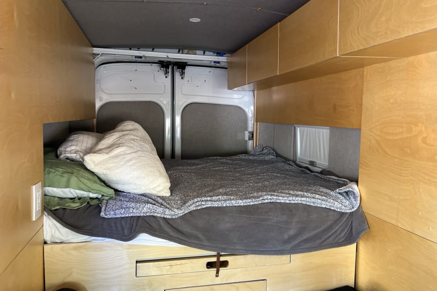 Explore this cozy RV rental's custom interior: a built-in bed with pillows, wooden cabinets, and rear doors.