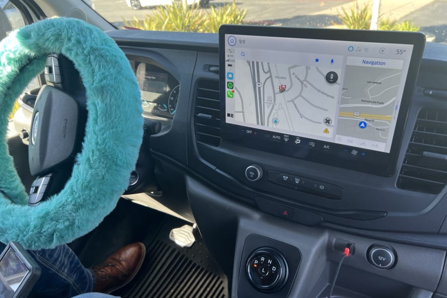 Inside a modern rv rental, a fluffy teal steering wheel cover contrasts with the large infotainment screen displaying navigation to Novato.