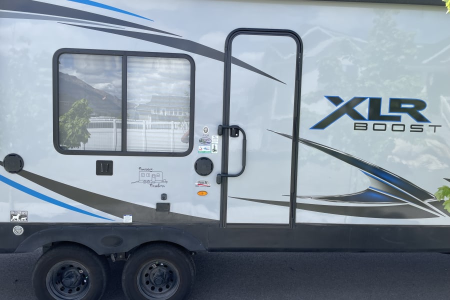 Side view of a grey XLR Boost travel trailer, ideal for rv rental. Features blue & black graphics, window, door, and wheels.
