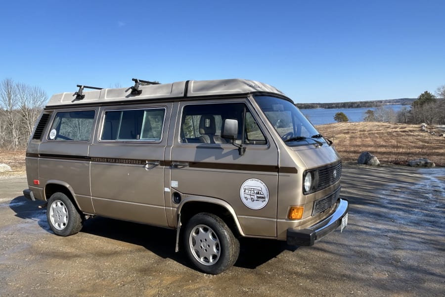 Tan vintage VW camper van with pop-top, branded "Vintage Van Adventures", for RV rental by a lake.