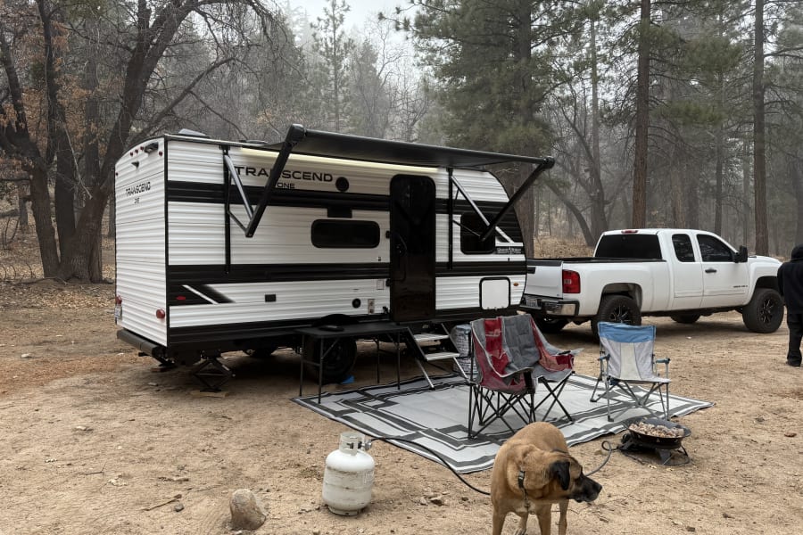 Cozy RV rental setup in a misty forest campsite, complete with chairs, fire pit, and a dog enjoying nature.